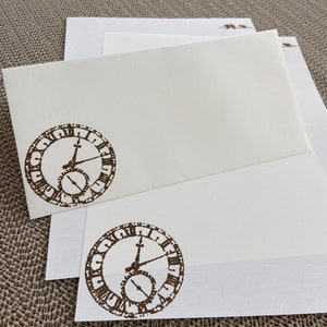 May include: Two white stationery note cards with brown clock face designs. The clock face has Roman numerals and a brown hand.