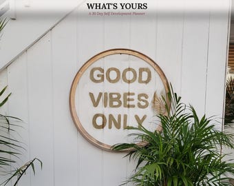 May include: A circular wooden wall sign featuring the text GOOD VIBES ONLY in tan letters. Above the sign, text reads ANDREA UNLOCKED, FOCUS ON WHAT'S YOURS, and A 30 Day Self Development Planner. The sign is mounted on a white vertical paneled wall.