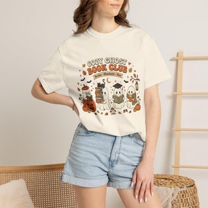 May include: An off-white short-sleeve t-shirt featuring a graphic print with the text "COZY GHOST BOOK CLUB" and "Books Blankets Boo". The design includes four cartoon ghosts, one inside a pumpkin, one holding books, one wearing a graduation cap, and one with a scarf, surrounded by autumn leaves, bats, and a lantern.