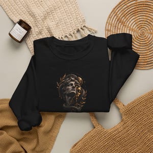 Könnte beinhalten: Ein schwarzes Langarm-Sweatshirt mit einem zentralen Grafikdruck einer klassischen Büste im Stein-Stil, die von einer Schlange umschlungen ist, akzentuiert durch goldene Blätter und ein kreisförmiges himmlisches Element vor dunklem Hintergrund.