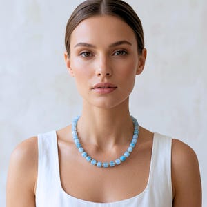 May include: A necklace featuring a series of light blue, cube-shaped beads alternating with small, round metallic spacers. The beads have a translucent, polished finish and are strung together to form a uniform, circular strand around the neck.