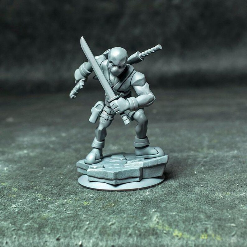 May include: A gray unpainted miniature figurine of a masked character in a crouching stance, holding a sword in one hand and a pistol in the other, with a second sword sheathed on the back, standing on a textured rocky base.
