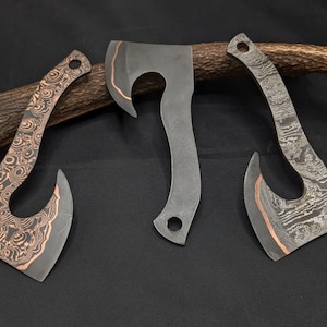 May include: Three metal hatchets with distinct finishes. The left hatchet features a copper and dark metal swirl pattern. The center hatchet is solid dark gray metal. The right hatchet displays a layered Damascus steel pattern with copper accents.