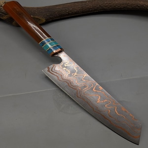 Peut inclure: Un couteau de cuisine avec une lame en acier Damas présentant des motifs ondulés de couleur cuivre. Le manche est en matériau brun lisse avec une virole décorative incrustée de segments turquoise et bleus, terminée par une virole en métal poli.