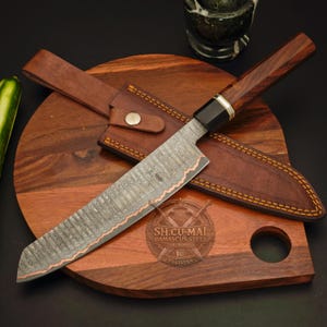 Peut inclure: Un couteau en acier Damas avec un tranchant cuivré et un manche en bois foncé, accompagné d'un étui en cuir marron avec des coutures jaunes. La lame présente un motif texturé et le texte SH.CU-MAI DAMASCUS STEEL ARTWORKS PAKISTAN.