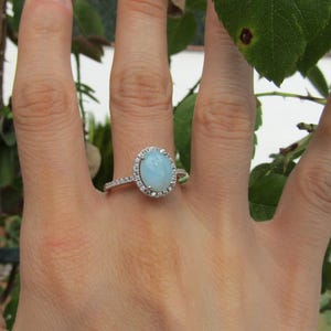 Puede incluir: Un anillo con una piedra central ovalada de color azul claro en cabujón rodeada por un halo de pequeñas piedras transparentes. La banda es fina y metálica, decorada con pequeñas piedras transparentes adicionales en los laterales.
