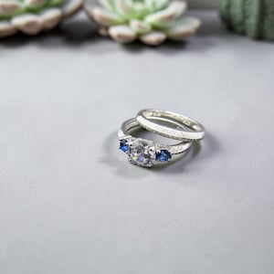 Puede incluir: Un conjunto de anillos de plata compuesto por una banda texturizada y un anillo principal con una gran piedra central transparente flanqueada por dos piedras azules más pequeñas. El metal tiene un acabado mate y granulado.