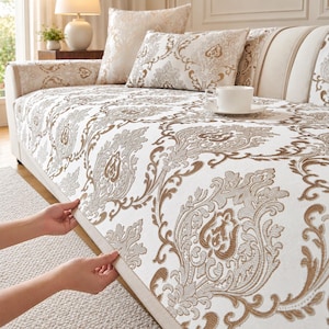 May include: A white sofa cover featuring a repeating brown and beige damask pattern. The fabric is draped over a light-colored sofa, which is accented with matching damask throw pillows and a plain white ceramic teacup on a saucer.