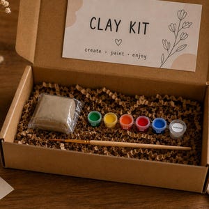 May include: A brown cardboard box containing a clay kit with a block of grey clay, a wooden paintbrush, and six small paint pots in green, yellow, orange, red, blue, and white. The box lid features the text CLAY KIT, create, paint, enjoy.