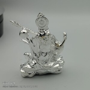 May include: A metallic silver-toned figurine depicting a seated deity holding a stringed musical instrument. The figure features intricate embossed details, a circular headpiece, and a textured base, all rendered in a reflective, polished finish.
