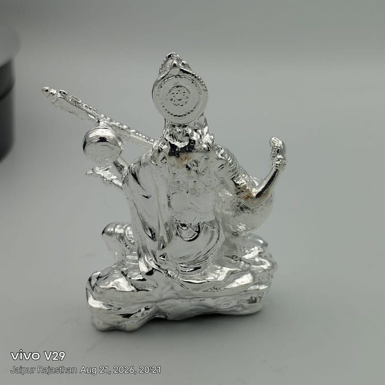 May include: A metallic silver-toned figurine depicting a seated deity holding a stringed musical instrument. The figure features intricate embossed details, a circular headpiece, and a textured base, all rendered in a reflective, polished finish.