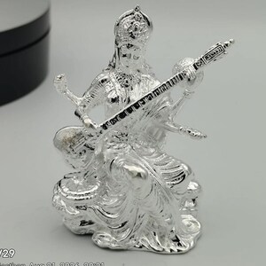 May include: A shiny silver-toned figurine depicting a seated deity wearing an ornate crown and traditional attire. The figure holds a long, stringed musical instrument known as a veena across the lap, with four arms visible in a detailed metallic finish.