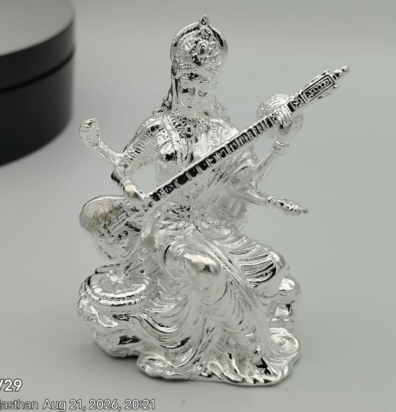 May include: A shiny silver-toned figurine depicting a seated deity wearing an ornate crown and traditional attire. The figure holds a long, stringed musical instrument known as a veena across the lap, with four arms visible in a detailed metallic finish.