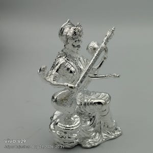 May include: A polished silver-toned figurine depicting a seated deity with four arms, holding a stringed musical instrument known as a veena. The figure wears an ornate headdress and traditional attire, captured in a reflective metallic finish.