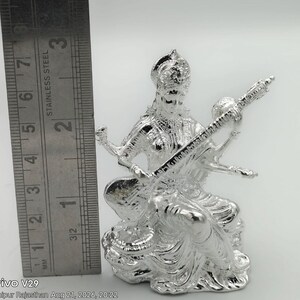 May include: A shiny silver-toned figurine of the goddess Saraswati playing a veena, positioned next to a stainless steel ruler showing a height of approximately 3.25 inches. The figure features intricate detailing on the clothing and jewelry.