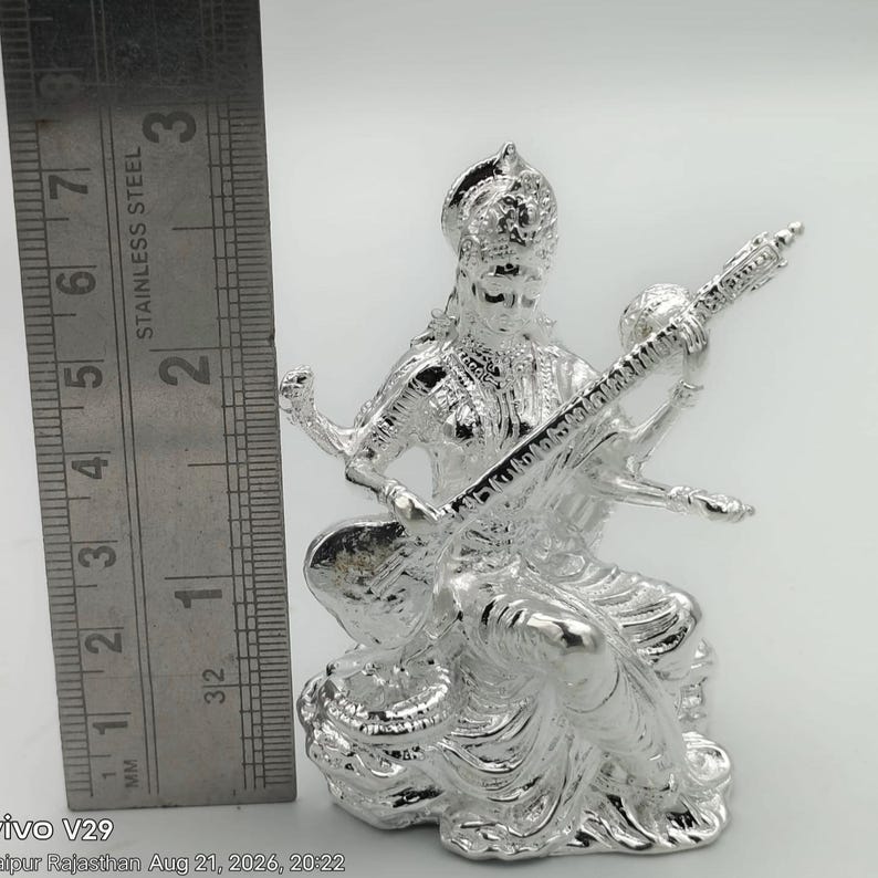May include: A shiny silver-toned figurine of the goddess Saraswati playing a veena, positioned next to a stainless steel ruler showing a height of approximately 3.25 inches. The figure features intricate detailing on the clothing and jewelry.