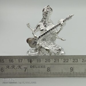 May include: A silver-colored figurine of a seated deity playing a stringed instrument, positioned behind a metal ruler showing measurements in inches, with the text A.R.K DELUXE visible on the ruler surface, capturing a detailed metallic finish.