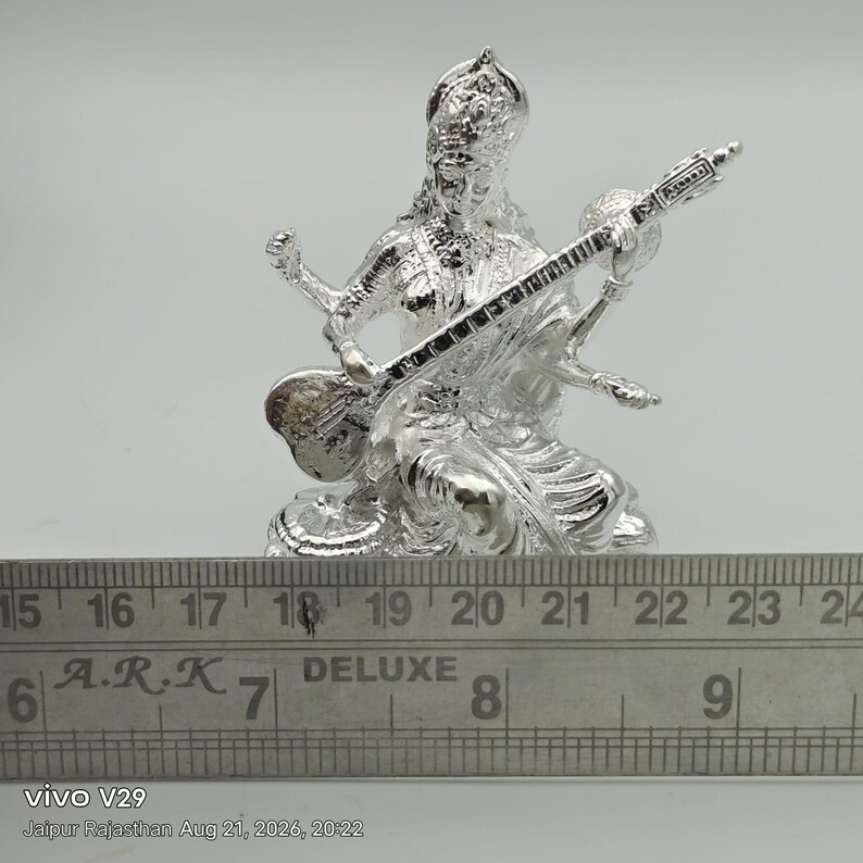May include: A silver-colored figurine of a seated deity playing a stringed instrument, positioned behind a metal ruler showing measurements in inches, with the text A.R.K DELUXE visible on the ruler surface, capturing a detailed metallic finish.
