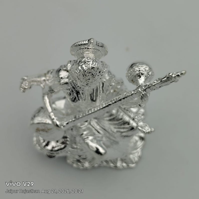 May include: A metallic silver-colored figurine depicting a seated deity holding a long, slender stringed musical instrument. The figure features intricate embossed details, a prominent headdress, and flowing garments, all rendered in a reflective, polished finish.