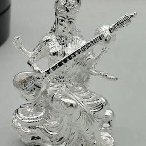 May include: A metallic silver-toned figurine depicting a seated deity with four arms, wearing an ornate crown and traditional garments. The figure holds a stringed musical instrument, a veena, across the body, with detailed carvings visible.