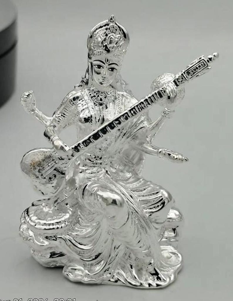 May include: A metallic silver-toned figurine depicting a seated deity with four arms, wearing an ornate crown and traditional garments. The figure holds a stringed musical instrument, a veena, across the body, with detailed carvings visible.