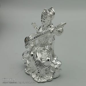 May include: A shiny silver-toned figurine depicting a seated deity playing a stringed veena instrument. The figure wears an ornate crown and traditional draped clothing with intricate detailing, captured in a reflective metallic finish.