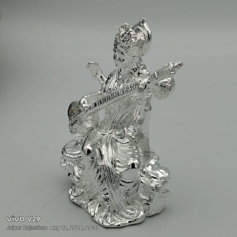 May include: A shiny silver-toned figurine depicting a seated deity playing a stringed veena instrument. The figure wears an ornate crown and traditional draped clothing with intricate detailing, captured in a reflective metallic finish.