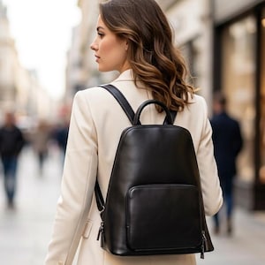 May include: A sleek black leather backpack featuring a smooth, rounded top design, a top carry handle, and a large rectangular front pocket with a zip closure. The backpack has adjustable shoulder straps and is worn over a light beige blazer.