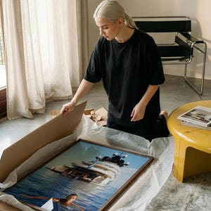 May include: A rectangular framed wall art print featuring a photographic image of a large white yacht anchored in deep blue water with a person swimming in the foreground. The artwork is encased in a dark brown wooden frame and rests in a cardboard box.