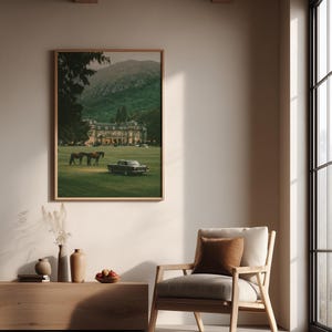 May include: A framed wall art print depicting a large estate with a green lawn, two horses, and a vintage car, displayed above a wooden console table with vases and a bowl of fruit, next to a light-colored wooden armchair with a brown cushion.