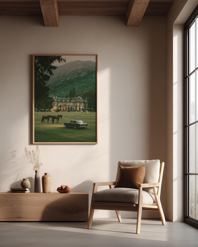 May include: A framed wall art print depicting a large estate with a green lawn, two horses, and a vintage car, displayed above a wooden console table with vases and a bowl of fruit, next to a light-colored wooden armchair with a brown cushion.