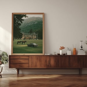 May include: A framed vertical wall art print featuring a landscape scene with a large manor house, two horses grazing on a green lawn, and a vintage black car parked in the foreground, all set against a backdrop of a forested mountain range.