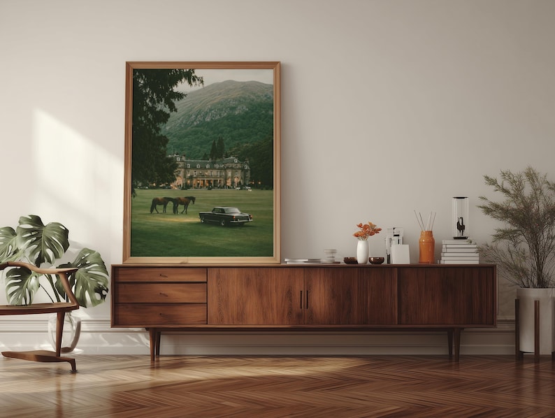 May include: A framed vertical wall art print featuring a landscape scene with a large manor house, two horses grazing on a green lawn, and a vintage black car parked in the foreground, all set against a backdrop of a forested mountain range.