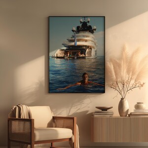 May include: A framed wall art print featuring a large white luxury yacht anchored in deep blue ocean waters with a person swimming in the foreground. The artwork is displayed in a room with a beige armchair, a wooden console table, and a vase of pampas grass.