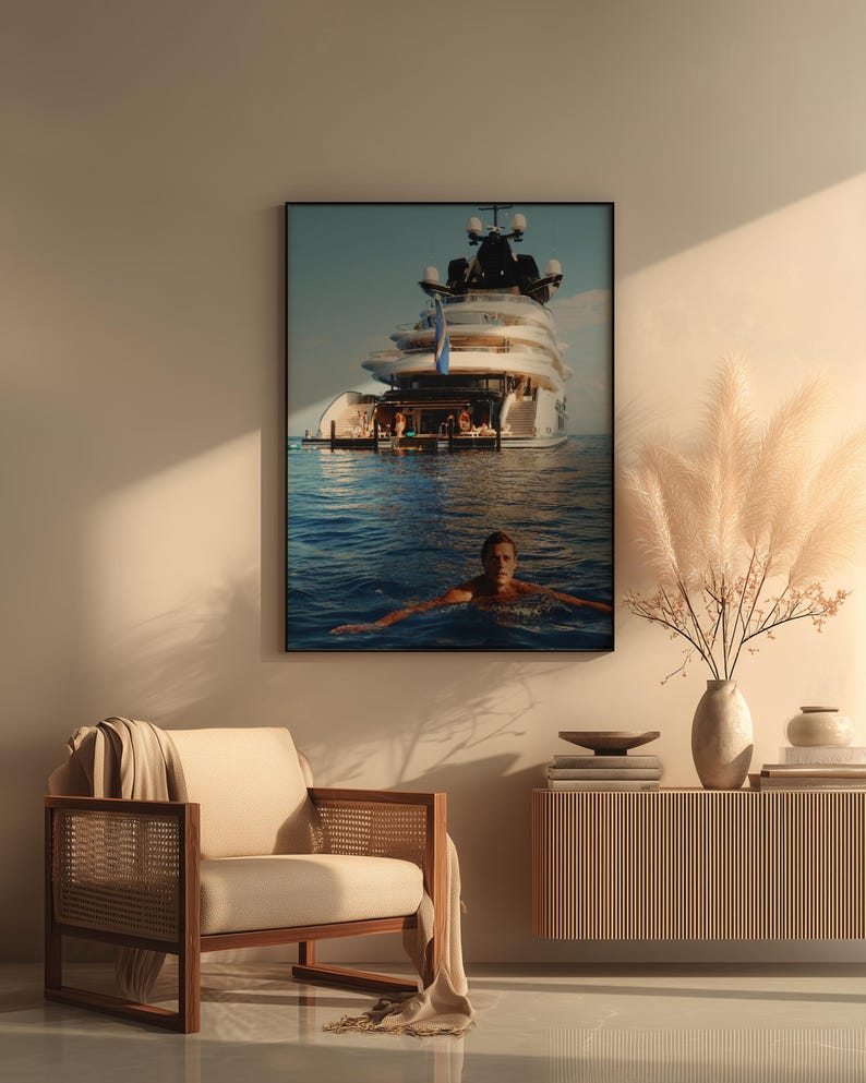May include: A framed wall art print featuring a large white luxury yacht anchored in deep blue ocean waters with a person swimming in the foreground. The artwork is displayed in a room with a beige armchair, a wooden console table, and a vase of pampas grass.