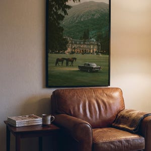 May include: A framed wall art print featuring a landscape scene with a large estate, two horses, and a vintage car on a grassy lawn in front of a mountain. The print is displayed in a black frame hanging on a wall above a brown leather armchair.