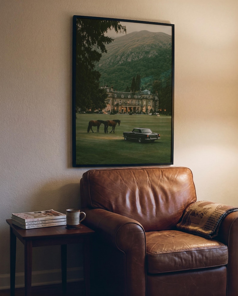 May include: A framed wall art print featuring a landscape scene with a large estate, two horses, and a vintage car on a grassy lawn in front of a mountain. The print is displayed in a black frame hanging on a wall above a brown leather armchair.