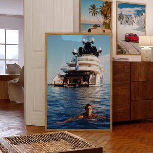 May include: A framed vertical wall art print featuring a large white luxury yacht floating on deep blue ocean water, with a person swimming in the foreground. The yacht has multiple decks and a blue flag, set against a clear sky with a small cloud.