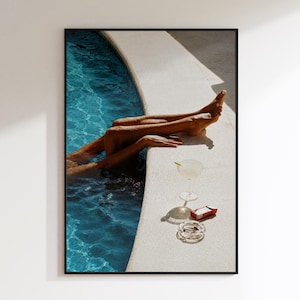 Könnte beinhalten: Ein gerahmter Wandkunstdruck, der ein Paar Beine am Rand eines Swimmingpools mit blauem Wasser zeigt. Auf dem hellen Beckenrand stehen ein klares Stielglas für Cocktails, eine kleine rote rechteckige Schachtel und ein runder Glasaschenbecher.