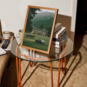 May include: A framed print featuring a landscape photograph of a large estate with a green lawn, two horses, and a dark vintage car, set against a mountain backdrop. The frame is made of light-colored wood and is displayed on a glass-topped table.