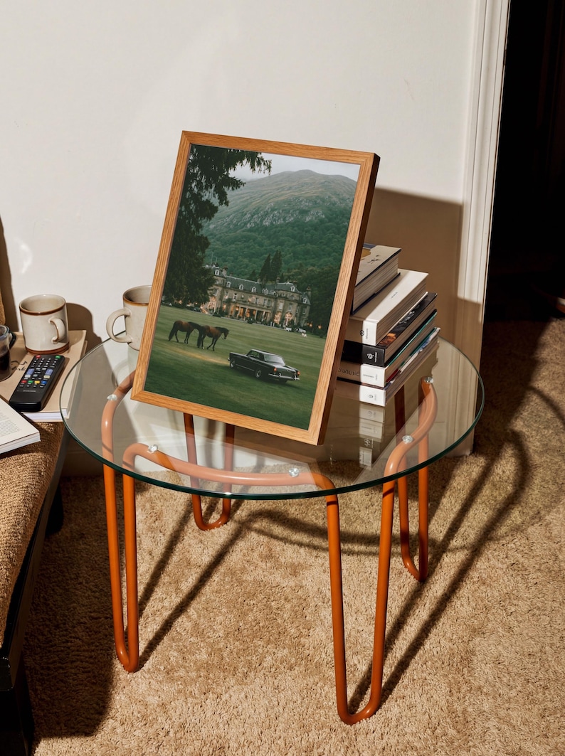 May include: A framed print featuring a landscape photograph of a large estate with a green lawn, two horses, and a dark vintage car, set against a mountain backdrop. The frame is made of light-colored wood and is displayed on a glass-topped table.