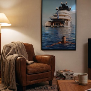 May include: A framed wall art print featuring a large white yacht anchored in blue water with a person swimming in the foreground. The artwork is displayed in a black frame on a wall above a brown leather armchair draped with a beige knit throw blanket.
