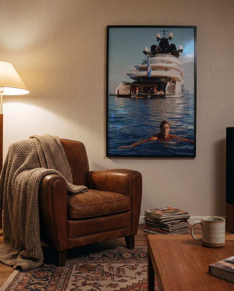 May include: A framed wall art print featuring a large white yacht anchored in blue water with a person swimming in the foreground. The artwork is displayed in a black frame on a wall above a brown leather armchair draped with a beige knit throw blanket.