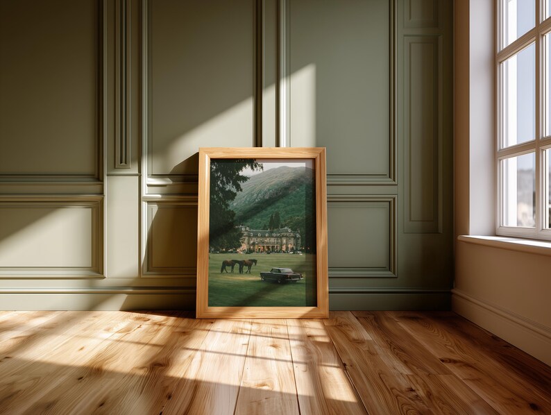 May include: A rectangular wooden picture frame containing a print of a large estate building situated on a grassy lawn with two horses and a vintage car. The frame is light brown with a natural wood grain finish and a simple, clean-lined border.