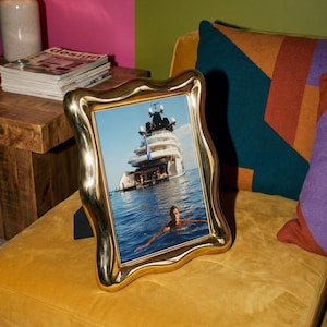 May include: A vertical rectangular photo frame with a polished gold-tone finish and a wavy, organic border. The frame contains a photograph of a large white yacht floating on blue ocean water with a person swimming in the foreground.