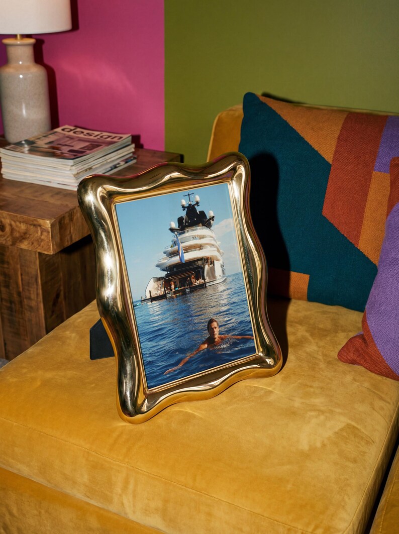May include: A vertical rectangular photo frame with a polished gold-tone finish and a wavy, organic border. The frame contains a photograph of a large white yacht floating on blue ocean water with a person swimming in the foreground.