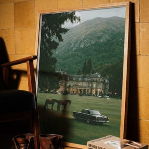 May include: A framed landscape photograph featuring a large manor house, two horses, and a vintage car on a green lawn with a mountain backdrop. The print is housed in a light wood frame and displayed alongside brown leather boots and stacked books.