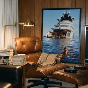 May include: A brown leather lounge chair with a matching ottoman, featuring a tufted back and seat. A gray throw blanket rests on the seat. Beside the chair is a stack of books and a brass lamp. A large framed photograph of a yacht is displayed.
