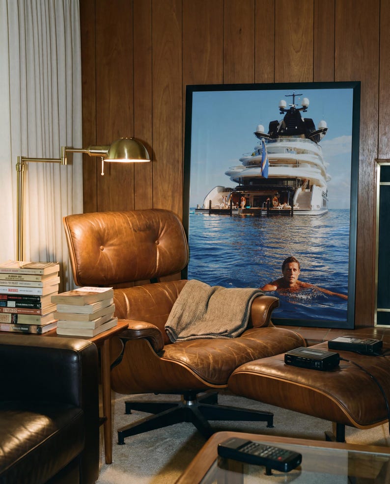May include: A brown leather lounge chair with a matching ottoman, featuring a tufted back and seat. A gray throw blanket rests on the seat. Beside the chair is a stack of books and a brass lamp. A large framed photograph of a yacht is displayed.