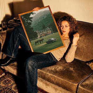 May include: A rectangular framed print featuring a landscape scene with a large manor house, two horses grazing on a green lawn, and a vintage car parked on the grass, all set against a backdrop of rolling hills and a dense forest.